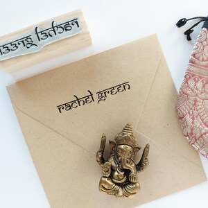 May include: A tan paper envelope with the name rachel green printed in black, a wooden rubber stamp with non-Latin script, a small bronze-colored Ganesha figurine, and a pink and gold patterned fabric drawstring pouch with a black top.