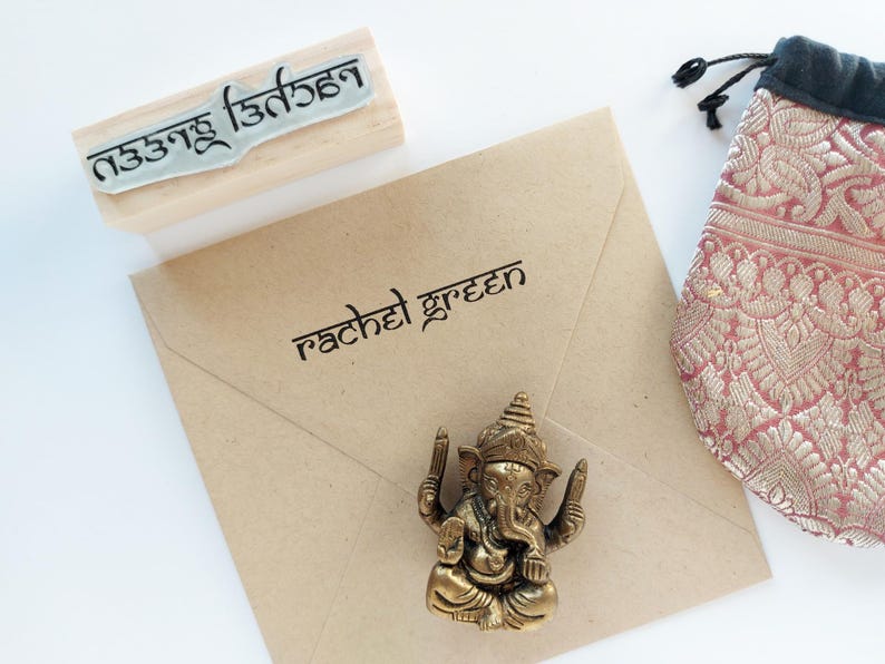 May include: A tan paper envelope with the name rachel green printed in black, a wooden rubber stamp with non-Latin script, a small bronze-colored Ganesha figurine, and a pink and gold patterned fabric drawstring pouch with a black top.