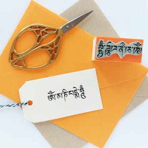 May include: A pair of gold-toned scissors with an intricate cutout handle design, an orange rubber stamp featuring Tibetan script, and a white rectangular paper tag with black Tibetan text and a black and white twine string, all on orange envelopes.