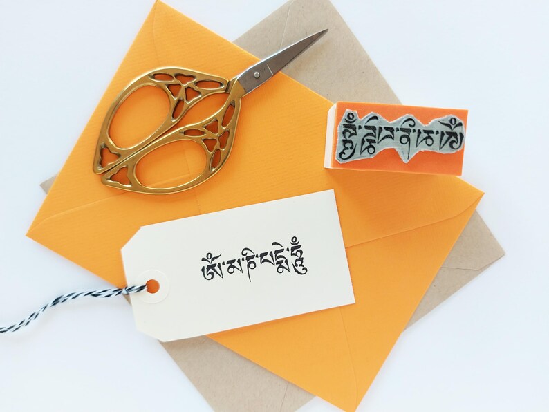 May include: A pair of gold-toned scissors with an intricate cutout handle design, an orange rubber stamp featuring Tibetan script, and a white rectangular paper tag with black Tibetan text and a black and white twine string, all on orange envelopes.