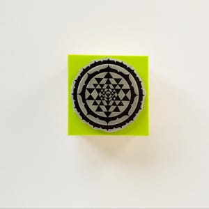 May include: A round, gray stamp with a geometric design, resting on a bright green square. The stamp features a detailed pattern of triangles and other shapes. The background is plain white.