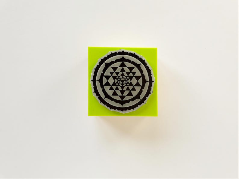 May include: A round, gray stamp with a geometric design, resting on a bright green square. The stamp features a detailed pattern of triangles and other shapes. The background is plain white.