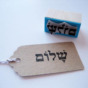 May include: A rectangular rubber stamp with a light wood handle and a bright blue base, featuring a black Hebrew inscription. Beside it lies a rectangular kraft paper tag with the same Hebrew word stamped in black ink.