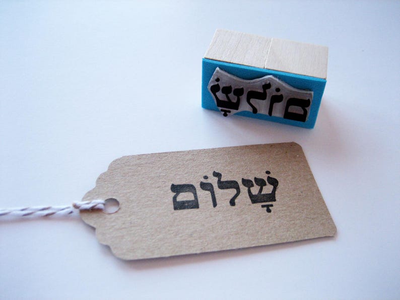 May include: A rectangular rubber stamp with a light wood handle and a bright blue base, featuring a black Hebrew inscription. Beside it lies a rectangular kraft paper tag with the same Hebrew word stamped in black ink.