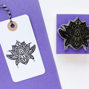 May include: A square rubber stamp with a black lotus flower design, positioned next to a white rectangular paper tag featuring the same black lotus print. The tag is attached to a purple card with a black and white twisted string.