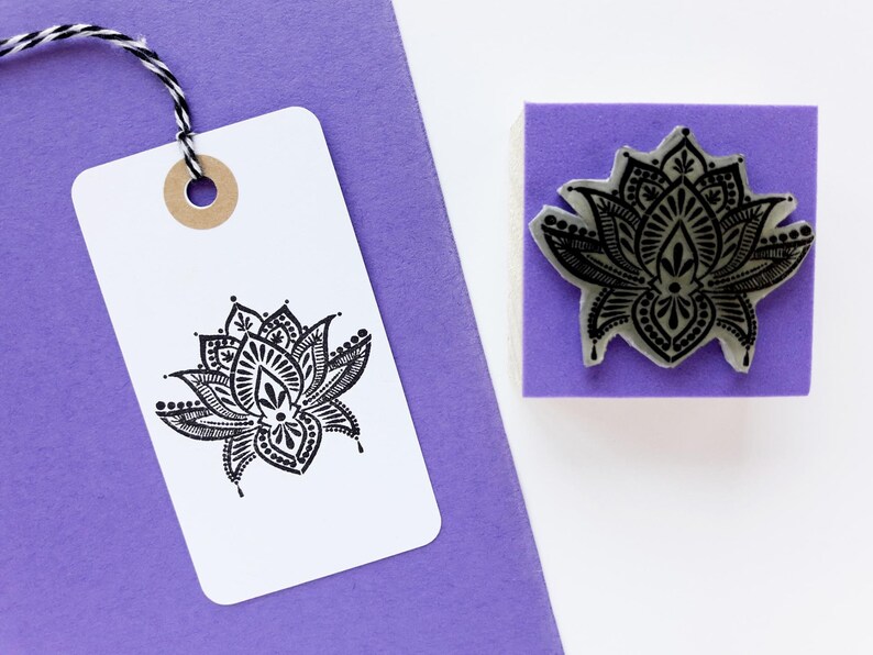 May include: A square rubber stamp with a black lotus flower design, positioned next to a white rectangular paper tag featuring the same black lotus print. The tag is attached to a purple card with a black and white twisted string.