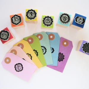 May include: A set of seven square wooden stamps with colorful foam tops, each featuring a unique black chakra symbol. Below the stamps are seven rectangular paper tags in various pastel colors, each stamped with a matching black chakra design.