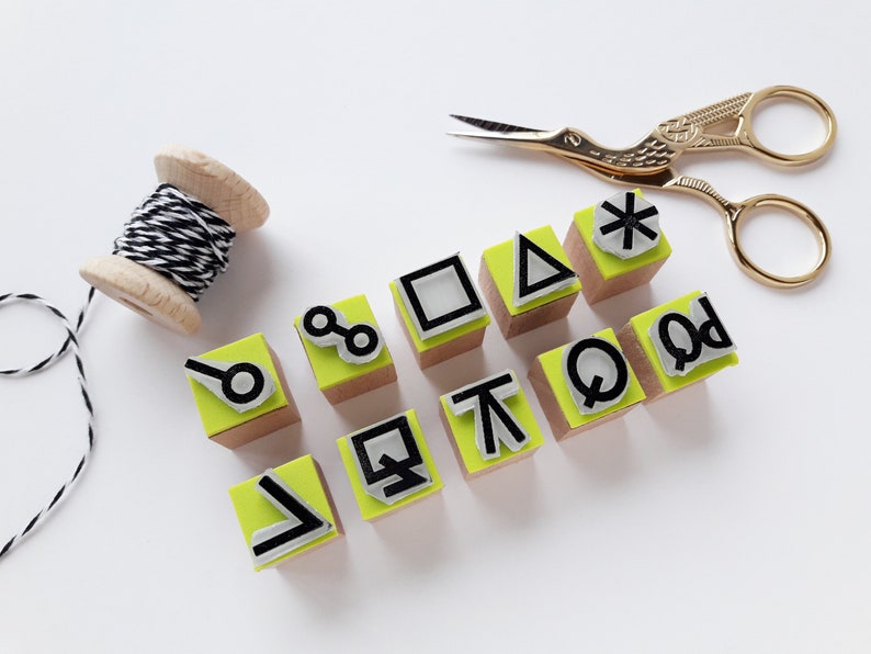 Op de afbeelding: Een set van tien houten stempels met verschillende geometrische vormen en symbolen, waaronder een cirkel, een vierkant, een driehoek, een sterretje en een paar abstracte vormen. De stempels zijn geverfd in een heldere geelgroene kleur en hebben zwarte inkt.