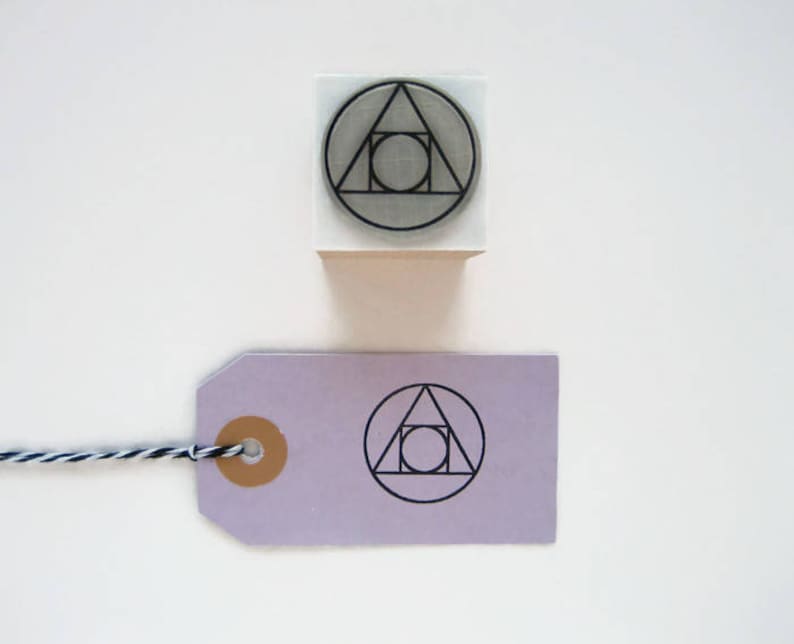 May include: A white rubber stamp with a black geometric design of a circle, triangle, and square. The stamp is on a white surface next to a purple tag with the same design.