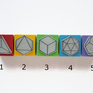 May include: Five wooden blocks, each with a different geometric shape outlined in black. The blocks are arranged in a row from left to right, with the colors red, yellow, green, blue, and purple. The shapes are a triangle, a circle, a cube, a hexagon, and an octagon. The numbers 1 through 5 are printed below the blocks.