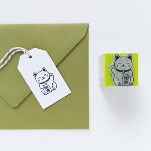 May include: A Maneki Neko stamp and tag set. The stamp features a gray cat with a raised paw on a green block. A white tag with a matching cat illustration is attached to a green envelope with twine.