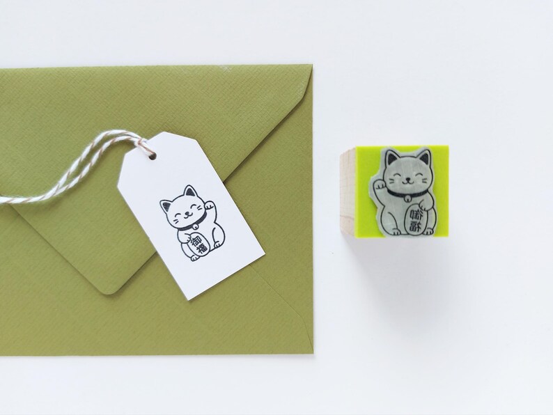 May include: A Maneki Neko stamp and tag set. The stamp features a gray cat with a raised paw on a green block. A white tag with a matching cat illustration is attached to a green envelope with twine.