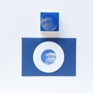 May include: A blue rubber stamp with a wave design, a blue envelope, and a white circular card with a wave design. The stamp is on a blue block. The wave design is in silver and blue. The items are on a white background.
