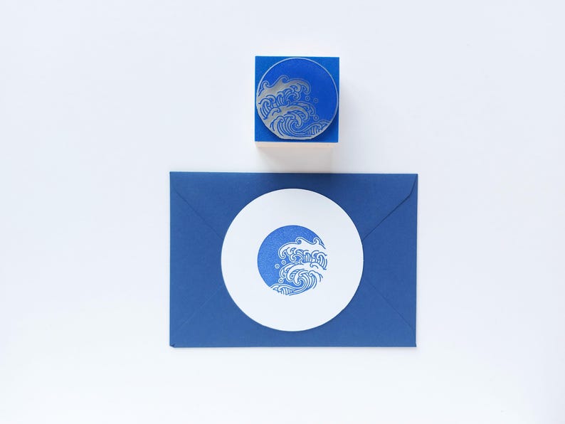May include: A blue rubber stamp with a wave design, a blue envelope, and a white circular card with a wave design. The stamp is on a blue block. The wave design is in silver and blue. The items are on a white background.
