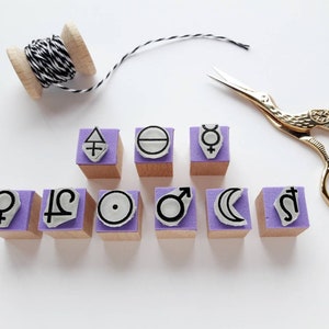 May include: Set of ten purple and white rubber stamps with astrological symbols, including the sun, moon, venus, mars, mercury, jupiter, saturn, and the zodiac signs for libra and aquarius. The stamps are arranged in two rows on a white surface.