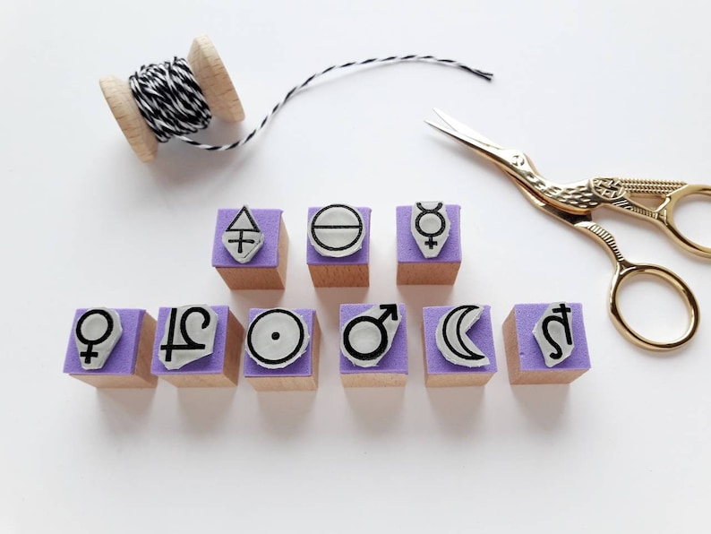 May include: Set of ten purple and white rubber stamps with astrological symbols, including the sun, moon, venus, mars, mercury, jupiter, saturn, and the zodiac signs for libra and aquarius. The stamps are arranged in two rows on a white surface.
