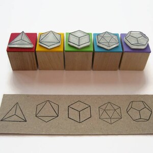 May include: Set of six wooden stamps with different geometric shapes. The stamps are in various colors, including red, yellow, green, blue, purple, and black. The shapes include a pyramid, a cube, a diamond, a hexagon, and a dodecahedron.