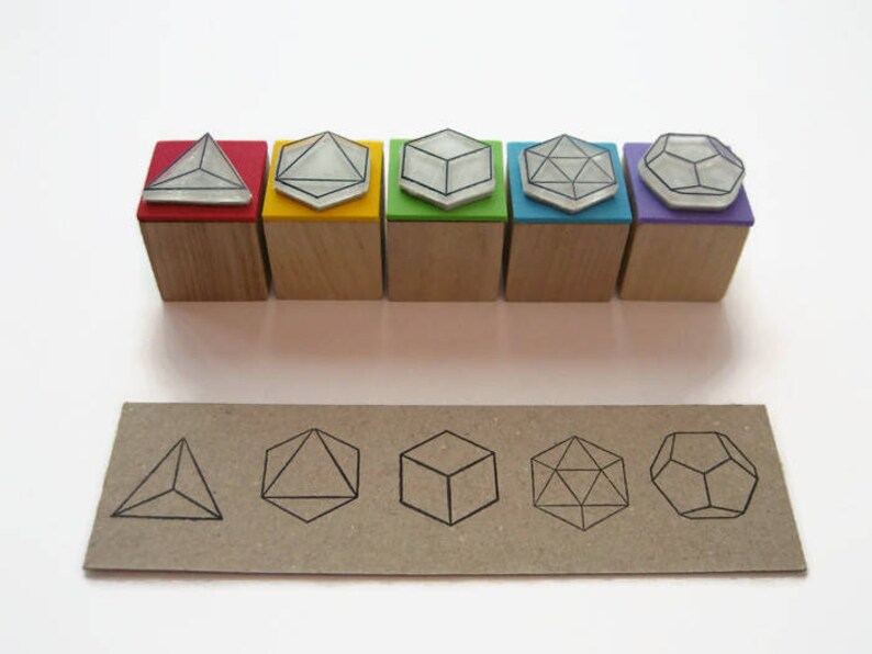 May include: Set of six wooden stamps with different geometric shapes. The stamps are in various colors, including red, yellow, green, blue, purple, and black. The shapes include a pyramid, a cube, a diamond, a hexagon, and a dodecahedron.