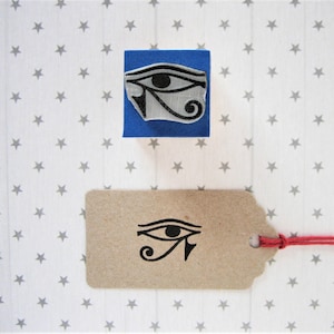 Op de afbeelding: Een blauwe rubberen stempel met een zwart-witte afbeelding van het oog van Horus. De stempel staat op een witte achtergrond met grijze sterren. Het oog van Horus is een symbool van bescherming en goede gezondheid in de oude Egyptische mythologie.