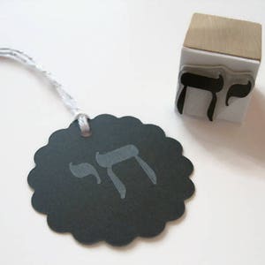 May include: A black scalloped circular paper tag with a silver string and the Hebrew letter Chai printed in gray. Beside it sits a square wooden rubber stamp featuring the same Hebrew letter Chai in black ink on a white rubber base.