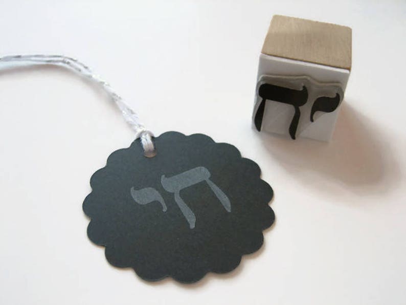 May include: A black scalloped circular paper tag with a silver string and the Hebrew letter Chai printed in gray. Beside it sits a square wooden rubber stamp featuring the same Hebrew letter Chai in black ink on a white rubber base.