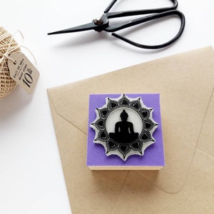 May include: Purple rubber stamp featuring a black silhouette of a meditating Buddha within a circular mandala design.