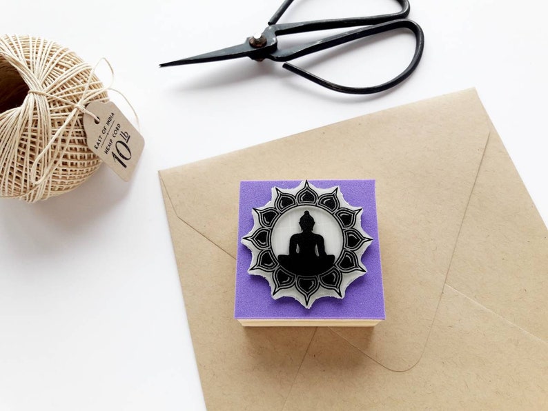 May include: Purple rubber stamp featuring a black silhouette of a meditating Buddha within a circular mandala design.