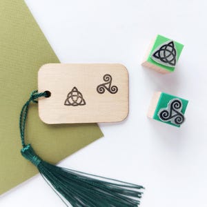 Op de afbeelding: Houten label en stempels met Keltische knoopontwerpen. Het label heeft een groene kwast. Er zijn ook twee stempels met groene bases en Keltische knoopontwerpen. De achtergrond omvat groen papier en een wit oppervlak.