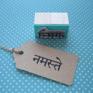 May include: A rectangular rubber stamp with a light wood handle and a teal base, featuring a raised black rubber design of the Devanagari script for Namaste. Beside it lies a rectangular brown paper tag with the word Namaste printed in black.
