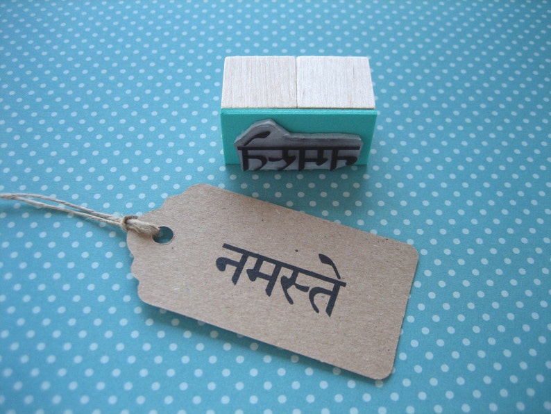 May include: A rectangular rubber stamp with a light wood handle and a teal base, featuring a raised black rubber design of the Devanagari script for Namaste. Beside it lies a rectangular brown paper tag with the word Namaste printed in black.