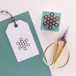 Op de afbeelding: Een wit cadeau-etiket met een zwart sneeuwvlokkenontwerp, een kleine houten stempel met een sneeuwvlokkenontwerp, een gouden schaar en een tealblauwe achtergrond.