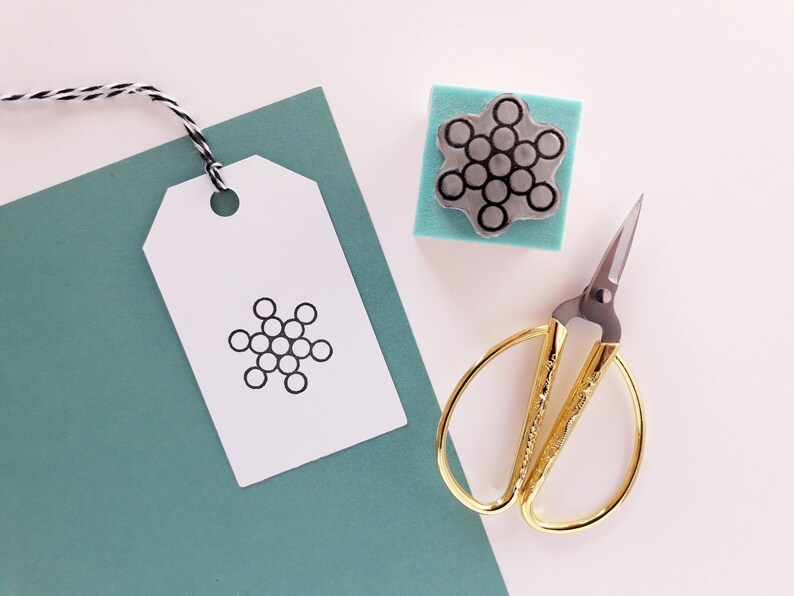 Op de afbeelding: Een wit cadeau-etiket met een zwart sneeuwvlokkenontwerp, een kleine houten stempel met een sneeuwvlokkenontwerp, een gouden schaar en een tealblauwe achtergrond.