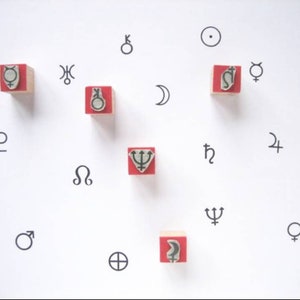 Op de afbeelding: Een wit oppervlak met rode houten blokken met verschillende astrologische symbolen, waaronder de zon, de maan, planeten en sterrenbeelden. De symbolen zijn in zwart en wit gedrukt.