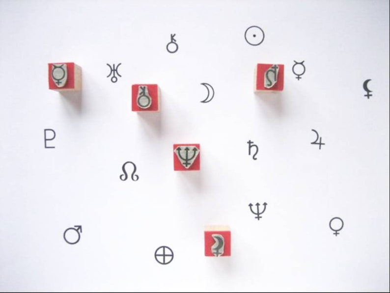 Op de afbeelding: Een wit oppervlak met rode houten blokken met verschillende astrologische symbolen, waaronder de zon, de maan, planeten en sterrenbeelden. De symbolen zijn in zwart en wit gedrukt.