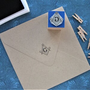 May include: A brown envelope with a stamped Masonic symbol, a blue stamp, and a black ink pad. The stamp features a compass and square design with the letter "G" in the center. Several small wooden clothespins are scattered around the envelope.