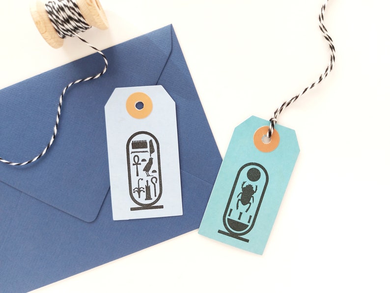 May include: Two blue and turquoise gift tags with black and white string. The tags feature black and white illustrations of Egyptian hieroglyphs. One tag has an image of a scarab beetle, the other has an image of an ankh.