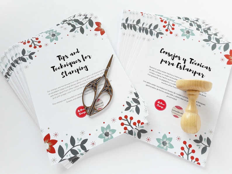 Op de afbeelding: Een stapel witte kaarten met een bloemenpatroon en de tekst "Tips and Techniques for Stamping" in zwart. Een stapel witte kaarten met een bloemenpatroon en de tekst "Consejos y T&eacute;cnicas para Estampar" in zwart. Een zilveren schaar en een houten stempel liggen op de kaarten.