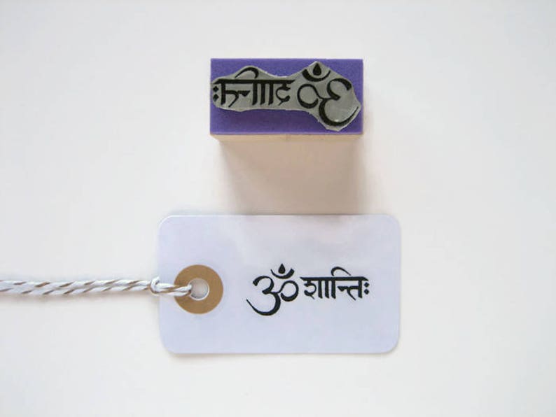 Puede incluir: Un sello de goma morado con la palabra s&aacute;nscrita "Om" y las palabras hindi "Shanti" en tinta negra. El sello est&aacute; sobre una superficie blanca y una etiqueta blanca con las palabras "Om Shanti" en tinta negra est&aacute; al lado.