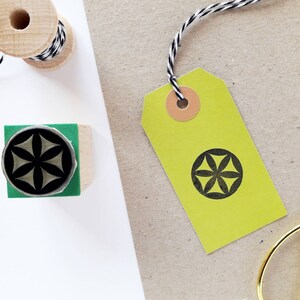 Op de afbeelding: Een geel cadeau-etiket met een geometrisch ontwerp in zwart en wit. Het etiket is vastgebonden met touw en heeft een bruine papieren cirkel bevestigd.