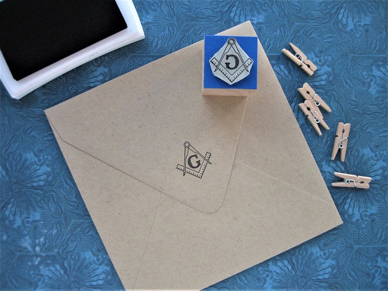 May include: A brown envelope with a black and white image of a compass and square with the letter "G" in the center. The envelope is on a blue patterned surface with wooden clothespins.