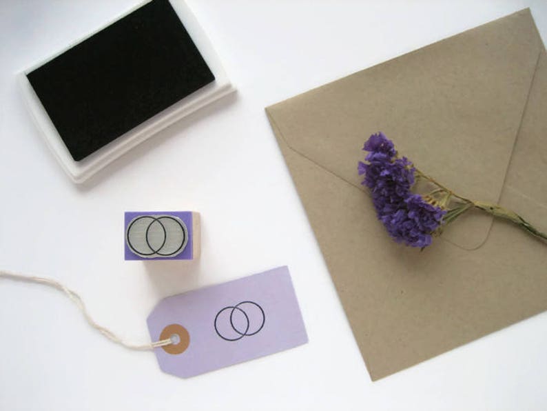 Op de afbeelding: Een houten rubberen stempel met twee overlappende cirkels in een lichtpaarse lijst. De stempel ligt op een wit oppervlak naast een paarse tag met een vergelijkbaar ontwerp en een kleine bruine cirkel.
