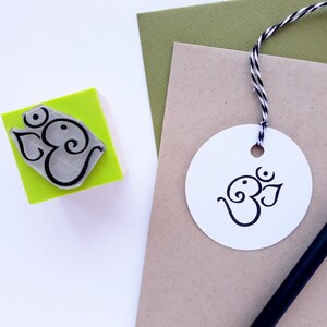 May include: A square lime green rubber stamp featuring a black Om symbol, alongside a white circular paper tag with the same black Om symbol attached by black and white twine, resting on tan and olive green paper with a black pencil nearby.