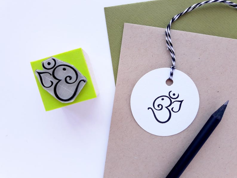 May include: A square lime green rubber stamp featuring a black Om symbol, alongside a white circular paper tag with the same black Om symbol attached by black and white twine, resting on tan and olive green paper with a black pencil nearby.