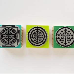 May include: Three square stamps with wooden bases and colored tops in teal, lime green, and forest green. Each stamp features a black Sri Yantra geometric design printed on a circular or square silver-toned plate attached to the top surface.