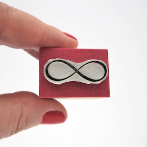 May include: A rectangular rubber stamp featuring a black infinity symbol design centered on a pink foam base. The stamp has a light-colored wooden block backing, providing a sturdy grip for the user while applying ink to paper or other surfaces.