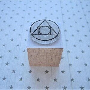 May include: A wooden block stamp with a clear, round, geometric design. The design features a triangle with a circle inside.