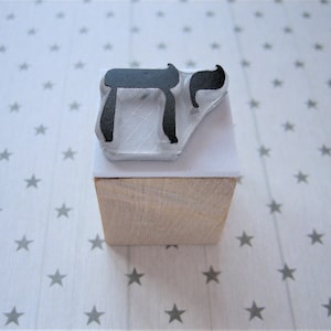 May include: A square wooden rubber stamp featuring the black Hebrew letter Chai on a white foam cushion. The stamp has a light-colored natural wood block base and a clear acrylic top layer that holds the black rubber design of the letter.