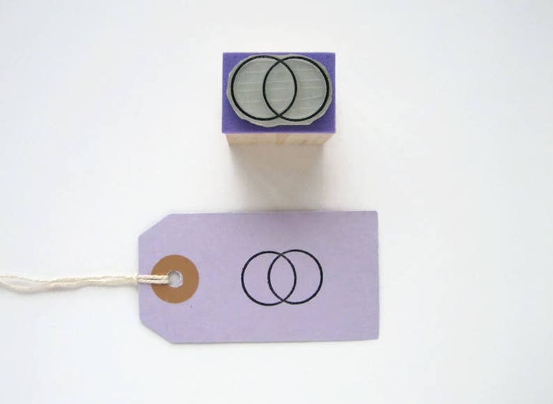 Op de afbeelding: Een houten stempel met een paarse basis en een wit en grijs ontwerp van twee overlappende cirkels. De stempel wordt gebruikt om een ontwerp van twee overlappende cirkels op een paars label te maken.