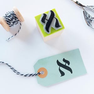 May include: A wooden spool of black and white baker's twine, a square wooden rubber stamp with a black letter H, and a light green paper gift tag with a black letter H, accompanied by a pair of silver metal scissors.