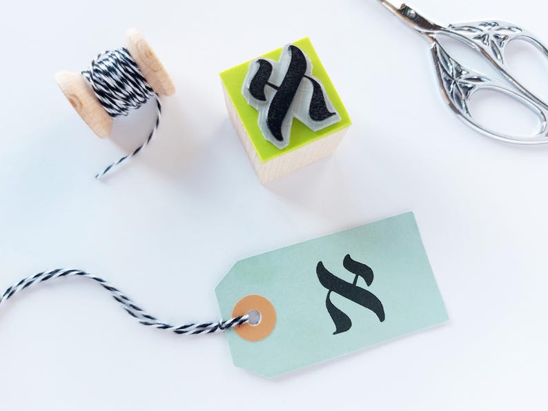 May include: A wooden spool of black and white baker's twine, a square wooden rubber stamp with a black letter H, and a light green paper gift tag with a black letter H, accompanied by a pair of silver metal scissors.